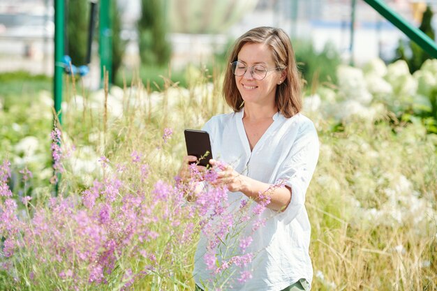 Wesoła dojrzała brunetka w okularach i białej koszuli robi zdjęcie fioletowych kwiatów na aparacie smartfona wśród innych roślin
