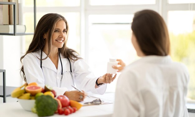 Wesoła dojrzała kaukaska pani doktor dietetyk w białym fartuchu doradza młodej kobiecie słoik witamin