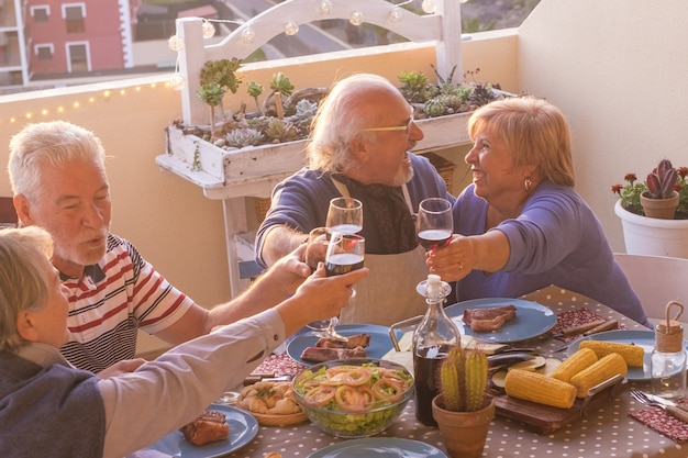 Wesoła dwie pary starszych świętują razem wznosząc toast na kieliszku wina na imprezie na tarasie. Osoby starsze bawiące się podczas posiłków na dachu. Starzy ludzie jedzący i pijący na imprezie na tarasie?