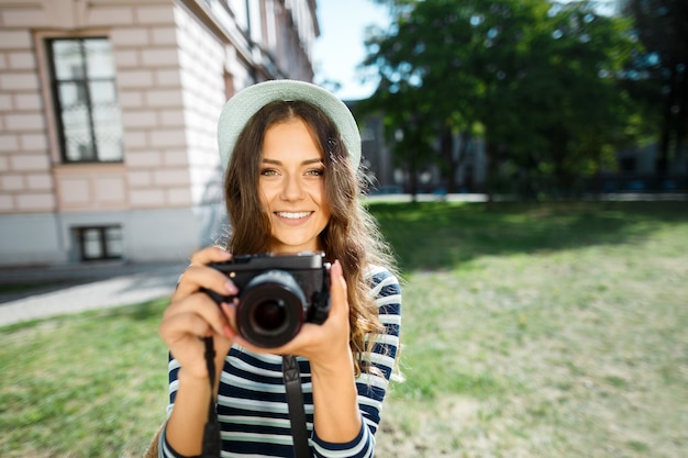 Wesoła dziewczyna turysta z kręconymi włosami w kapeluszu, trzymając aparat i uśmiechając się do starego miasta europejskiego, podróżowanie, Europa.