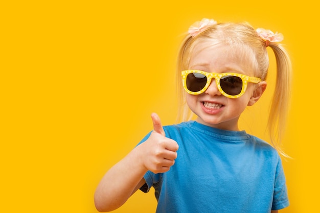 Wesoła dziewczynka w wieku przedszkolnym z blond włosami i dwoma kucykami nosi okulary przeciwsłoneczne Dziecko pokazuje aprobaty jak gest Kopiuj przestrzeń