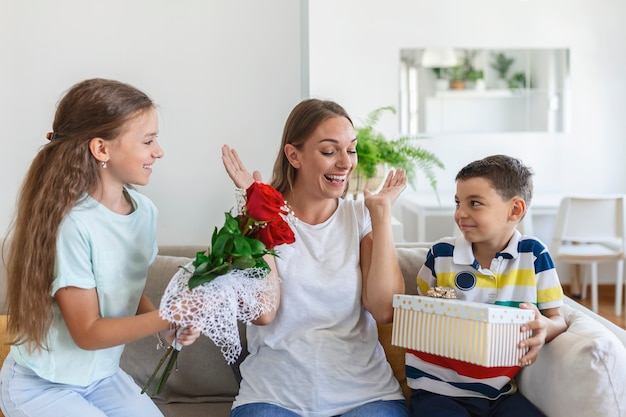 Wesoła dziewczynka z bukietem kwiatów róż i najmłodszy brat z pudełkiem uśmiecha się i gratuluje szczęśliwej mamy na dzień matki w domu. Szczęśliwego dnia Matki!