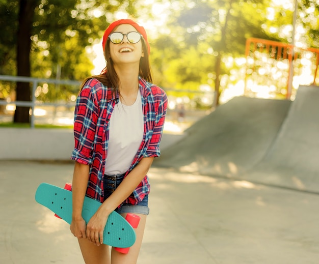Wesoła emocjonalna młoda kobieta ubrana w stylowe ubrania, trzymając w ręku deskorolkę w skateparku w jasny słoneczny dzień