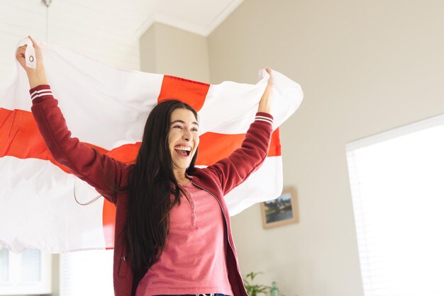 Wesoła kaukazyjska młoda kobieta podnosząca flagę Anglii i krzycząca podczas oglądania meczu w domu.