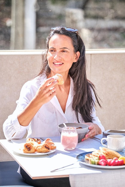 Wesoła kobieta jedząca pieczone croissanty na zdrowe śniadanie i odpoczywająca na tarasie kawiarni w słoneczny dzień