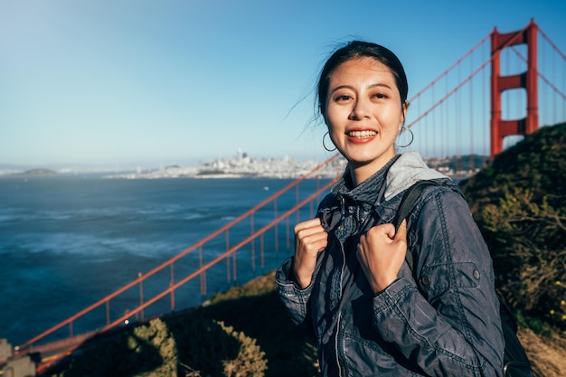 wesoła kobieta turysta śmiejąc się przez most Golden Gate w san francisco. piękna dziewczyna backpacker stojący na wzgórzach twarz aparat uśmiechający się w promieniach słońca. błękitne czyste niebo i woda oceanu morskiego.