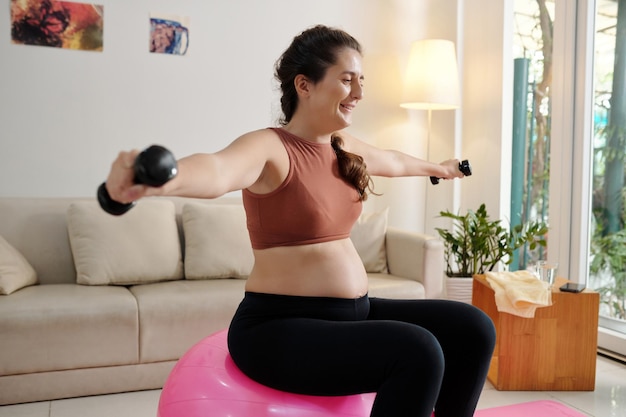Wesoła kobieta w ciąży siedzi na piłce fitness i robi ćwiczenia z małymi hantlami