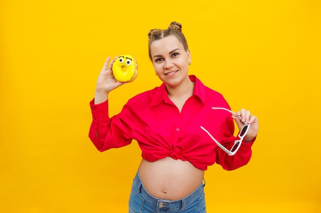 Wesoła kobieta w ciąży trzymająca w dłoni słodki żółty pączek na żółtym tle Oczekiwanie ciąży i macierzyństwa dziecka Koncepcja zdrowej i niezdrowej diety Fast food