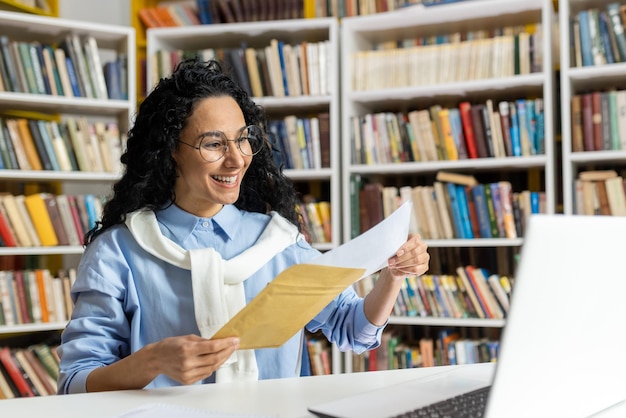 Zdjęcie wesoła kobieta w okularach czyta wiadomość pocztową z uśmiechem otoczona półkami z książkami w