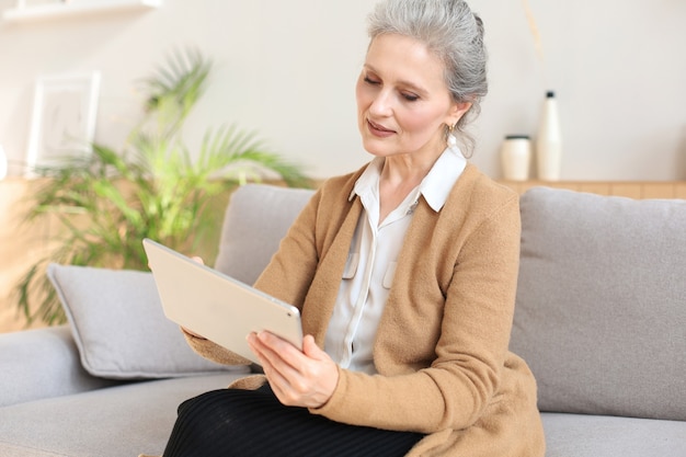 Wesoła kobieta w średnim wieku siedząca na kanapie, korzystająca z aplikacji na tablet komputerowy, patrząca na ekran, czytająca dobre wieści w sieciach społecznościowych, robiąca zakupy lub rozmawiająca online.