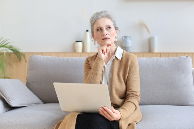 Wesoła kobieta w średnim wieku za pomocą laptopa siedząc na kanapie w domu.