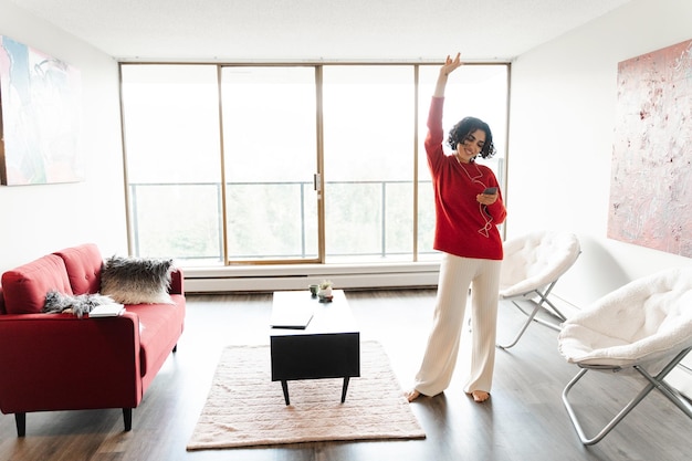 Wesoła kobieta z Bliskiego Wschodu trzymająca telefon komórkowy słuchająca muzyki i tańcząca w salonie Wesoła szczęśliwa kobieta spędzająca czas w domu