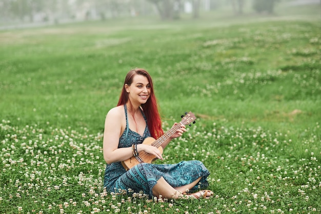 Wesoła kobieta z rudymi włosami grając na ukulele w parku, siedząc na trawie w letni dzień.