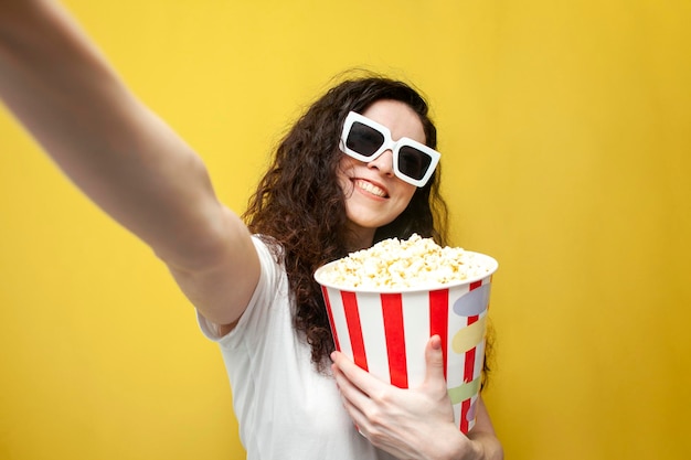 Wesoła kręcona dziewczyna w okularach 3d trzyma popcorn i bierze selfie na żółtym tle