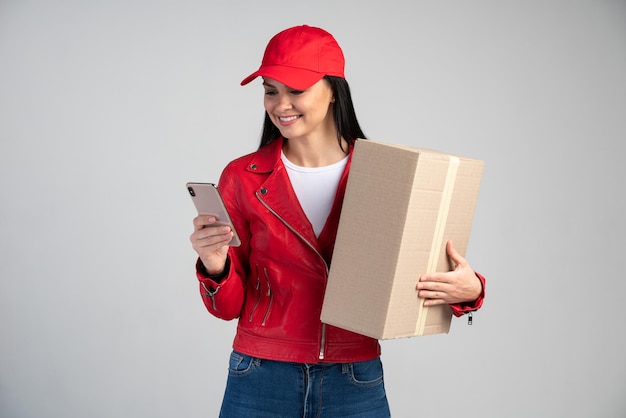 Wesoła kurierka trzymająca folder, dostarczająca zamówienie na adres, patrząca na ekran smartfona, ubrana w mundur. Kryty strzał studio na białym tle na szarym tle