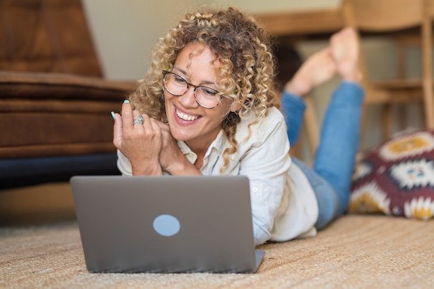 Wesoła ładna dorosła kobieta prowadzi wideorozmowę online z laptopem leżącym na podłodze w domu