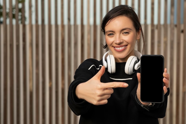 Wesoła, ładna kaukaska tysiącletnia kobieta w odzieży sportowej i słuchawkach pokazuje telefon z pustym ekranem