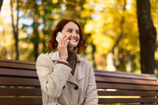 Wesoła, ładna kaukaska tysiącletnia kobieta z rudymi włosami w płaszczu przeciwdeszczowym dzwoniąca przez telefon cieszy się jesienią i spaceruje