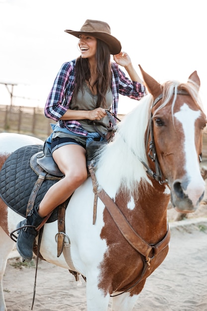 Wesoła ładna młoda kobieta cowgirl siedzi i jeździ konno w wiosce