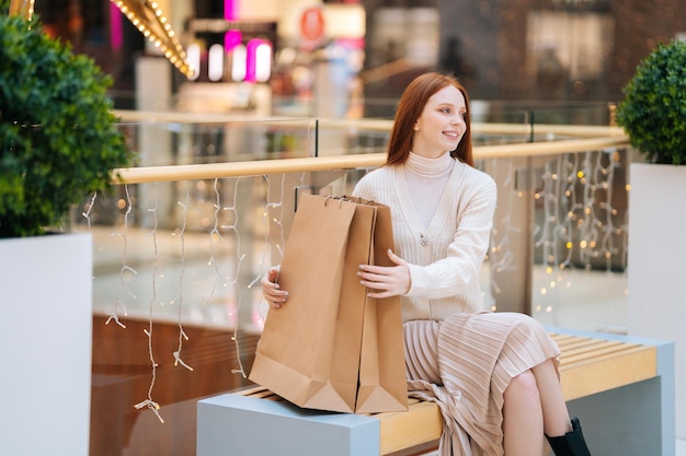 Wesoła ładna młoda kobieta siedzi na ławce z papierowymi torbami na zakupy w centrum handlowym z nowoczesnym wnętrzem