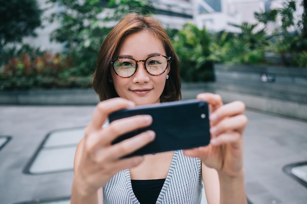 Wesoła, ładna młoda kobieta w stroju casual i okularach, robiąca selfie przez telefon komórkowy na zewnątrz