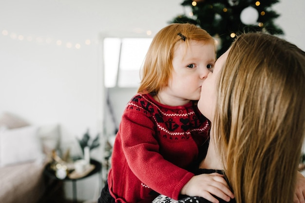 Wesoła mama całuje uroczą córeczkę w pobliżu choinki Wesołych Świąt i wesołych świąt Matka i małe dziecko bawią się i bawią razem w domu Zbliżenie