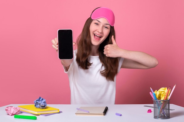 Wesoła miła dziewczyna trzyma w ręku inteligentny telefon pokazując pusty ekran na białym tle na różowym tle