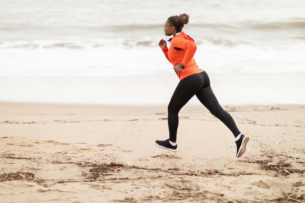 Wesoła młoda Afroamerykanka sportowiec w odzieży sportowej i słuchawkach biegnie na plaży morskiej na świeżym powietrzu