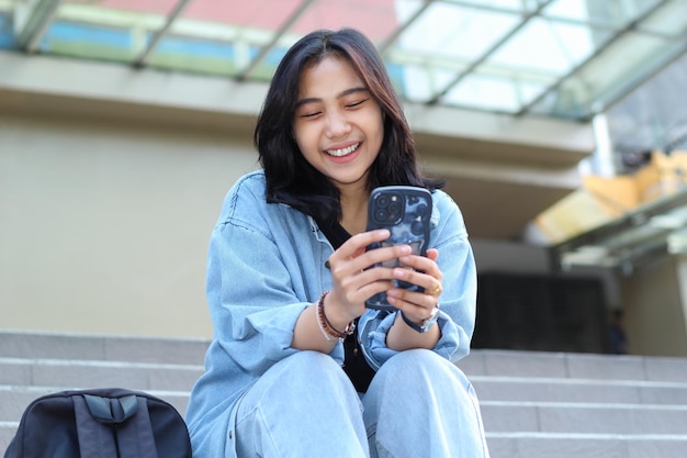 wesoła młoda Azjatka pracująca zdalnie śmieje się korzystając ze smartfona surfując w mediach społecznościowych aplikacja mobilna czatowanie wideo uzyskać zniżkę w sklepie internetowym siedząc na schodach miejskich na świeżym powietrzu
