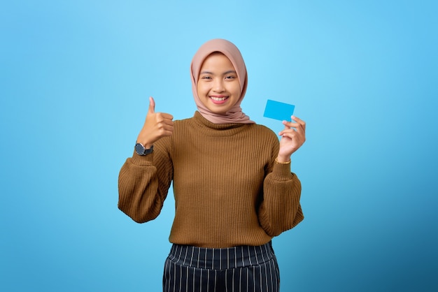 Wesoła młoda Azjatka trzymająca kartę kredytową i pokazująca kciuk w górę gest na niebieskim tle