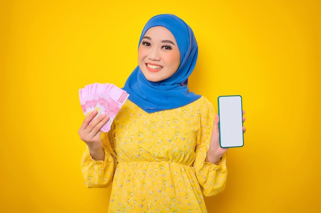 Wesoła młoda Azjatycka kobieta w casualowej sukience pokazująca pusty ekran telefon komórkowy i trzymająca banknoty pieniędzy na białym tle na żółtym tle