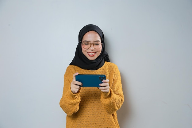 Wesoła młoda azjatycka muzułmanka w pomarańczowym swetrze i okularach grająca w grę na telefonie komórkowym