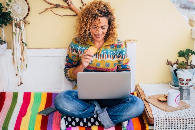 Wesoła młoda dorosła kobieta za pomocą komputera siedzi i wygodnie je ciastko Szczęśliwy styl życia w domu i surfowanie po sieci Współcześni ludzie pracują na laptopie w czasie wolnym online Alternatywne biuro