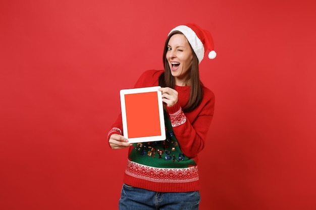 Wesoła młoda dziewczyna Santa w Boże Narodzenie kapelusz miga, przytrzymaj komputer typu tablet pc z pustym pustym ekranem na białym tle na czerwonym tle. Szczęśliwego nowego roku 2019 celebracja party wakacje koncepcja. Makieta miejsca na kopię.