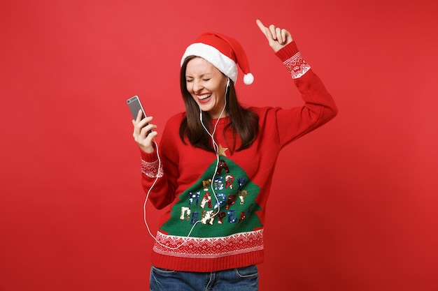 Wesoła młoda dziewczyna Santa ze słuchawkami, trzymając telefon komórkowy, słuchanie muzyki, rosnąca ręka, taniec na białym tle na czerwonym tle. Szczęśliwego nowego roku 2019 celebracja party wakacje koncepcja. Makieta miejsca na kopię.