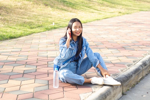 Wesoła młoda dziewczyna siedząca w parku w mieście trzyma telefon komórkowy z butelką wody umieszczoną na bocznej koncepcji stylu życia