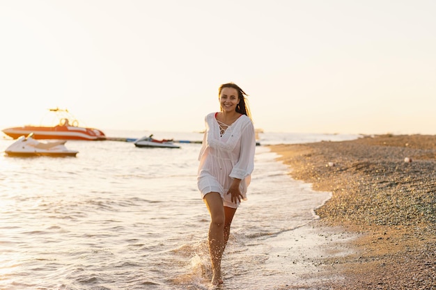 Zdjęcie wesoła młoda kobieta biegnie wzdłuż piaszczystej plaży o zachodzie słońca z łodziami na tle