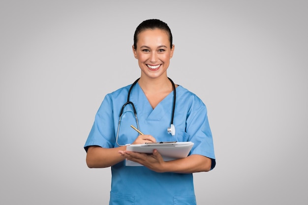 Wesoła młoda kobieta, lekarz, chirurg lub pielęgniarka w niebieskim mundurze, robiąca notatki na tablicy i uśmiechająca się