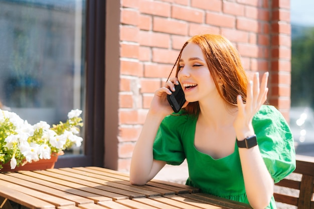 Wesoła młoda kobieta o rozmowie na telefon komórkowy siedzi przy stole na tarasie kawiarni na świeżym powietrzu w słoneczny letni dzień. Piękna ruda pani rozmawia smartfon siedząc w kawiarni w europejskim mieście