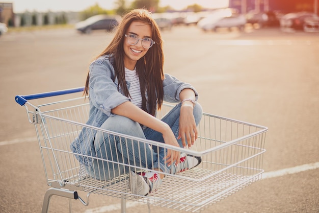Wesoła młoda kobieta siedzi w wózku na zakupy