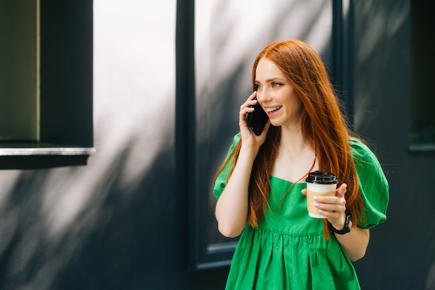 wesoła młoda kobieta trzymająca filiżankę kawy na wynos, rozmawiająca przez telefon komórkowy spacerująca po ulicy miasta