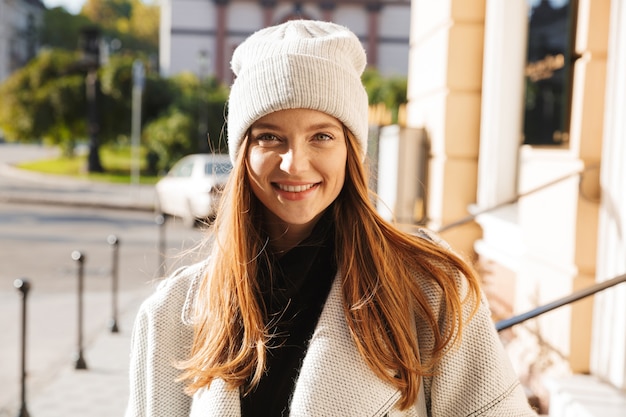 Wesoła młoda kobieta ubrana w jesienny płaszcz i kapelusz spaceru na świeżym powietrzu na ulicy miasta