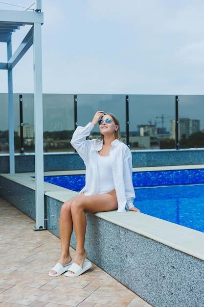 Wesoła młoda kobieta w białym stroju kąpielowym i białej koszuli siedzi przy basenie Kobieta w okularach przeciwsłonecznych i stroju kąpielowym Wakacje nad basenem Opalona dziewczyna