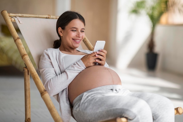 Wesoła młoda kobieta w ciąży za pomocą smartfona podczas relaksu w fotelu w domu