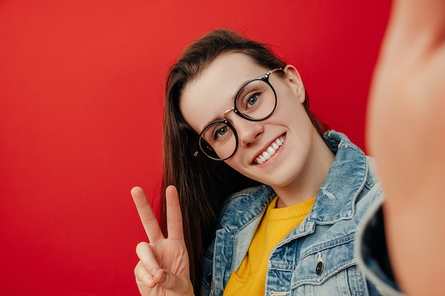 Wesoła młoda kobieta w okularach, robi gest pokoju, bierze selfie na smartfona, dobrze się bawi z komórkową, nosi dżinsową kurtkę, pozuje na czerwonym tle z pustą kopią