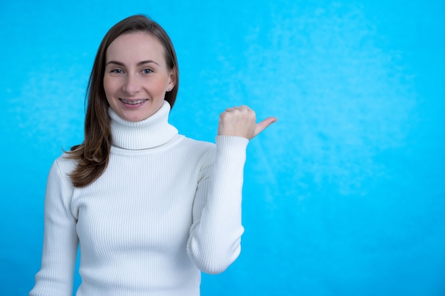 Wesoła młoda kobieta, wskazując kciukiem na bok, promuje nowy przedmiot, uśmiecha się przyjemnie, odizolowana na niebieskiej ścianie.