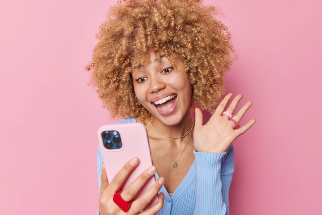Wesoła młoda kobieta z kręconymi włosami macha dłonią i wita przyjaciela online ma uśmiechy wideokonferencji z zębami używa nowoczesnego smartfona ubrana swobodnie, na białym tle na różowym tle Koncepcja technologii