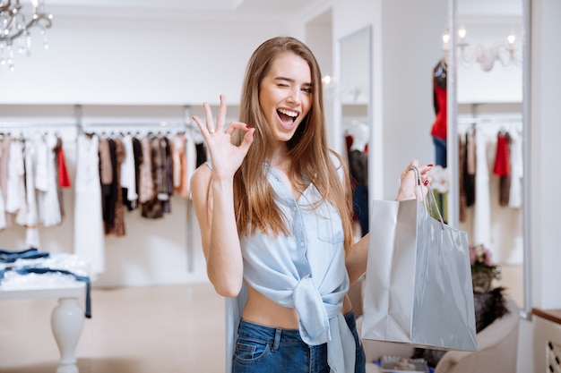Wesoła młoda kobieta z torbą na zakupy, mrugając i pokazując ok znak w sklepie odzieżowym