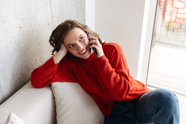 Wesoła młoda niedbale ubrana kobieta siedzi na kanapie w domu, rozmawiając przez telefon komórkowy