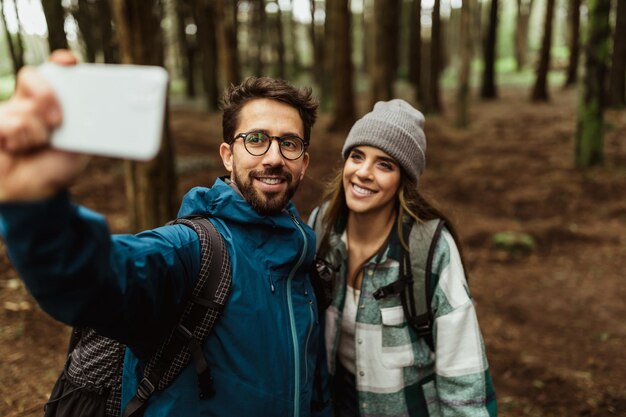 Wesoła młoda para białych w kurtkach w lesie robi selfie na smartfonie cieszy się wakacjami